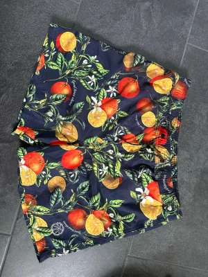Mönstrade badbyxor från Jack & Jones - Färgglada badbyxor från Jack & Jones med tropiskt mönster av apelsiner, citroner och gröna blad på mörkblå botten. Resår i midjan och innerbyxa i mesh för extra komfort. Perfekta för stranden eller poolen.