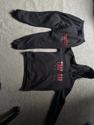 Svart hoodie Trapstar London - Säljer en svart hoodie från Trapstar London med röd och vit logga på bröstet och texten 'LONDON' i vitt. Hoodien har magficka och huva med dragsko. Perfekt för dig som gillar streetwear och vill ha en snygg och bekväm stil.