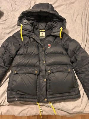Fjällräven Expedition down lite - Snygg svart dunjacka från Fjällräven med huva och gula dragskor. Modellen är Expedition down lite i storlek S. Jackan har två stora fickor med knappar framtill och klassisk Fjällräven-logga på bröstet. Perfekt för kalla vinterdagar tack vare det tjocka dunfodret och den rejäla designen. Använd en vinter så fortfarande i mycket bra skick! 💕