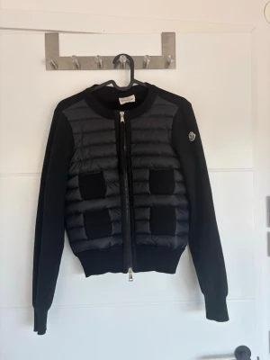 Svart cardiganjacka med quiltad front - Snygg svart cardiganjacka från Moncler med quiltad, vadderad front och två fickor. Ärmar och rygg i mjukt stickat material, dragkedja framtill och logga på ärmen. Perfekt för lager-på-lager och streetstyle. Har fått den i present så inte helt säker på äktheten men man kommer till monclers egna hemsida med qr koden 