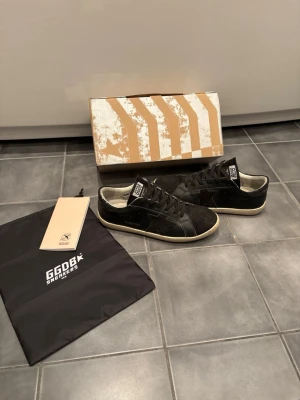 Golden goose skor - Säljer ett par svarta GGDB sneakers i en clean low-modell. Skorna är gjorda i en mix av mocka och läder med vita detaljer och vit sula. Klassisk stjärndesign på sidan och GGDB-logga på plösen. Perfekta för dig som gillar stilrena och trendiga sneakers. Storlek 42, hör av dig vid intresse mvh