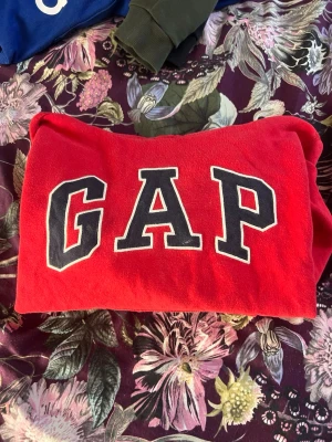 Röd hoodie från GAP med logga - Säljer en klassisk röd hoodie från GAP med stor svartvit logga framtill. Hoodien har en avslappnad passform och är tillverkad i mjukt material, perfekt för en chill och casual stil. Passar dig som gillar streetwear och vill ha något bekvämt.