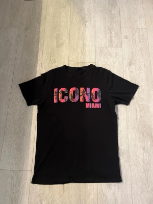 ICONO T-shirt M - Snygg svart t-shirt med färgglatt tryck där det står 'ICONO MIAMI' i rosa och lila toner med blommigt mönster i bokstäverna. Klassisk passform och rund hals. Perfekt för dig som vill sticka ut lite extra.