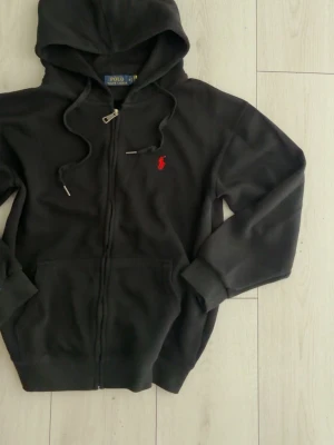 Svart hoodie från Polo Ralph Lauren - Snygg svart hoodie från Polo Ralph Lauren med dragkedja, huva och dragsnören. Klassisk röd logga broderad på bröstet och känguruficka framtill. Perfekt för en avslappnad och stilren look.