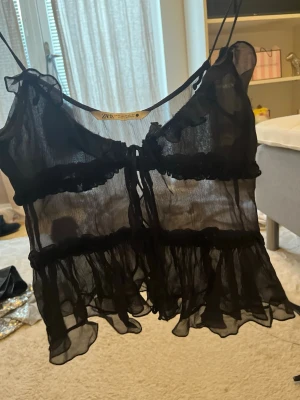 Svart transparent volanglinnetopp Zara - Svart transparent linnetopp från Zara med tunna axelband, volangdetaljer och knyt framtill. Toppen har en flowy passform och romantiska volanger längs kanterna, perfekt för lager på lager och trendiga looks.