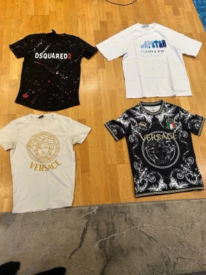 Fyra trendiga t-shirts från kända märken - 300KR STYCK! 1000KR FÖR ALLA! Fyra olika t-shirts: en svart DSQUARED2 med färgstänk och tryck, en vit Napstar med blått tryck, en vit Versace med guldtryck och en svart/vit Versace med detaljerat mönster och emblem. Alla har korta ärmar och är perfekta för dig som gillar streetwear och designerplagg.