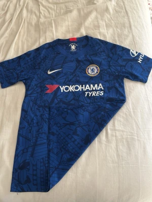 Chelsea FC matchtröja blå Nike - Chelsea FCs officiella matchtröja i blått med grafiskt mönster, klubbmärke på bröstet och Nike-logga. Tröjan har sponsortryck från Yokohama Tyres och Hyundai på ärmen. Tillverkad i lätt och ventilerande material, perfekt för fotbollsträning eller match. Tröjan är i storlek L (barnstorlek) och passar i ca 11-13 år. Tröjan är använd någon gång och är i väldigt bra skick.