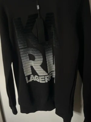 Svart Karl Lagerfeld hoodie - Svart hoodie från Karl Lagerfeld med stort, grått och vitt logotryck framtill. Tröjan har långa ärmar, ribbade muddar och en klassisk passform. Perfekt för dig som gillar streetwear och vill sticka ut med en snygg logga.
