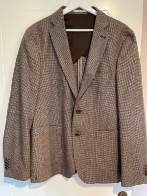 Brunmelerad kavaj med hundtandsmönster, strl 50 - Stilren kavaj från Pour i brunmelerat tyg med klassiskt hundtandsmönster. Tvåknäppt framtill, fickor med lock och knappar vid ärmslut. Perfekt för en dressad look och passar till både jeans och kostymbyxor.