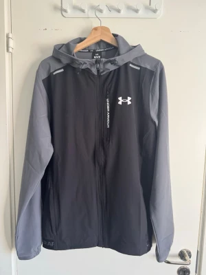 Under armour vanish woven jacket  - Snygg vindjacka från Under Armour i svart och grått med huva och hel dragkedja. Jackan har logga på bröstet, praktiska fickor och elastiska muddar. Perfekt för blåsiga dagar och sportiga outfits. Lappen finns kvar och kvitto finns. Som ny finns bevis på kvittot att den är en vecka gammal. Stolen M men passar S