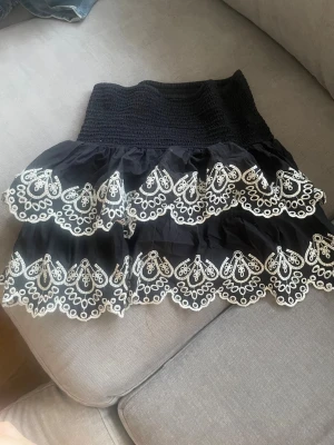 Svart kortkjol med vit brodyr - Supersöt svart kortkjol med bred smock i midjan och två volanger. Kjolen har vit broderad spetsdetalj längs kanterna som ger en romantisk känsla. Perfekt att styla med en enkel topp eller stilren tröja💕