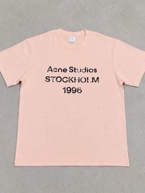 Ljusrosa Acne Studios t-shirt - Ljusrosa t-shirt från Acne Studios med svart tryck på bröstet där det står 'Acne Studios STOCKHOLM 1996'. Klassisk passform och rund halsringning. Perfekt för en avslappnad och trendig stil.