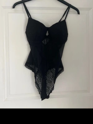 Svart spetsbody med tunna axelband - Snygg svart body i spets med tunna axelband och transparenta meshpartier. Bodyn har vackra spetsdetaljer och en feminin skärning som framhäver figuren. Perfekt för dig som vill ha något extra 