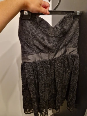Svart spetsklänning - Säljer en svart axelbandslös kortklänning med vacker spets och markerad midja med satinband. Klänningen har en omlottliknande överdel och volangig kjol. Perfekt för dig som vill ha en elegant och stilren look. Kan inte hitta någon storlek. Men uppskattar den till M.