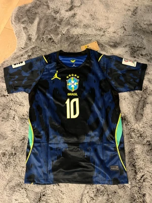 Brasilien Neymar Jr matchtröja  - Säljer en blå och svart Brasilien matchtröja med Neymar Jr och nummer 10 på ryggen. Tröjan har gula och turkosa detaljer, korta ärmar och officiella tryck framtill och bak. Perfekt för fotbollsfans som vill sticka ut på planen eller läktaren.