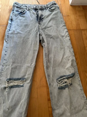 Ljusa raka/wide jeans med slitningar - Säljer ett par ljusblå raka/wide jeans med tydliga slitningar och hål vid knäna. Jeansen har fem fickor och klassisk dragkedja med knapp. Perfekta för en avslappnad och trendig streetstil. Materialet är denim i bomull.
