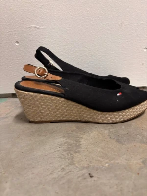 Svarta kilklackade sandaler Tommy Hilfiger - Snygga svarta sandaler med öppen tå och kilklack i flätad bast från Tommy Hilfiger. Skon har en justerbar rem med metallspänne och diskret logga på sidan. Ovandelen är i tyg och innersulan i brunt läderliknande material.