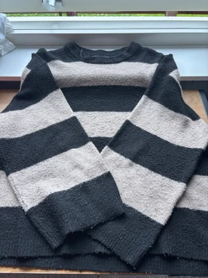 Randig svart och beige stickad tröja - Säljer en stickad tröja med breda svarta och beige ränder. Tröjan har rund halsringning, långa ärmar och är gjord i ett mjukt, lite luddigt material. Perfekt för dig som gillar en avslappnad och trendig look.
