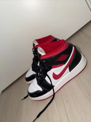 Air Jordan 1 Mid svart/röd/vit - Säljer ett par äkta Nike Air Jordan 1 Mid sneakers i svart, rött och vitt. Storlek 37,5. Klassisk design med svart läderöverdrag, vit bas och röda detaljer som swoosh och foder. Skorna har snörning och den ikoniska Air Jordan-loggan på sidan. Har bara använt dessa skor 1-2 gånger max, så de är i princip oanvända och nya. 