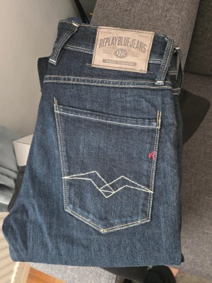 Mörkblå Replay jeans med kontrastsömmar - Säljer ett par mörkblå jeans från Replay med snygga kontrastsömmar och klassisk femficksmodell. Jeansen har en unik broderad detalj på bakfickan och Replay-logga på etiketten. Materialet är mjukt denim som sitter skönt och passar till många olika stilar.