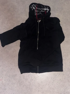 Svart hoodie med rutigt foder - Snygg svart hoodie med dragkedja och huva. Insidan av huvan och fodret har ett rutigt mönster i svart, vitt och rött som ger en cool kontrast. Hoodien har fickor framtill och ribbade muddar vid ärmar och nederkant. Perfekt för en avslappnad streetstyle.