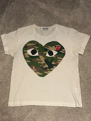 Vit Comme des Garçons PLAY t-shirt - Säljer en vit t-shirt från Comme des Garçons PLAY med ett stort hjärta i camouflage-mönster och ögon på bröstet. Liten röd hjärtdetalj med ögon i hörnet. Klassisk rund hals och korta ärmar. Perfekt statement-plagg för dig som gillar streetwear.