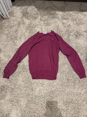 Mörklila rundhalsad tröja - En stilren mörklila tröja med rund halsringning och långa ärmar. Tröjan har ribbade muddar vid ärmslut och nederkant. Perfekt för dig som gillar en enkel men snygg look. Materialet ser ut att vara mjukt och tunt, troligen bomull eller bomullsblandning.