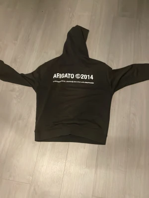 Svart hoodie med ARIGATO-tryck - Säljer en svart/grå hoodie med vit text 'ARIGATO ©2014' tryckt på både bröstet och ryggen. Hoodien har en klassisk huva och en stor magficka framtill. Perfekt för dig som gillar stilrena och moderna plagg med streetwear-vibe.