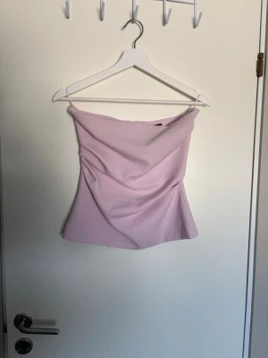 Ljuslila / rosa axelbandslös tubtopp - Säljer en snygg ljuslila tubtopp med draperad framsida. Toppen är axelbandslös och har en stilren, figurnära passform. Perfekt att styla med jeans eller kjol för en trendig look.