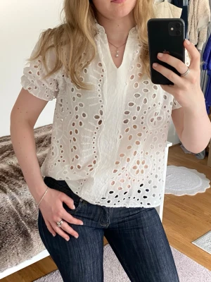 Vit broderad blus med kort ärm - Säljer en söt vit blus med broderade hålmönster och volangdetaljer vid ärmslut. Blusen har korta puffärmar och en liten ståkrage med v-ringning. Tillverkad i ett lätt och luftigt tyg, perfekt för sommaren🤍🤍🤍🤍🤍🤍