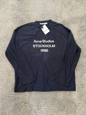 Mörkblå långärmad topp Acne Studios - Mörkblå långärmad topp från Acne Studios med vit text 'Acne Studios STOCKHOLM 1996' på bröstet. Klassisk rund halsringning och avslappnad passform. Perfekt för en stilren och trendig look.