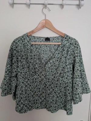 Grön blommig blus - Söt blus i ljusgrönt med blommigt mönster. Modellen har v-ringning, knappar framtill och vida trekvartsärmar. Tunt, luftigt tyg som känns perfekt till våren och sommaren!