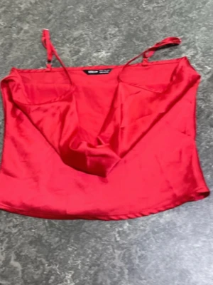 Röd satinlinnetopp med smala band - Snygg röd linnetopp i satin med tunna axelband och draperad urringning. Perfekt för dig som vill ha en trendig och stilren topp till sommaren. Materialet ger ett lyxigt intryck och färgen sticker verkligen ut.