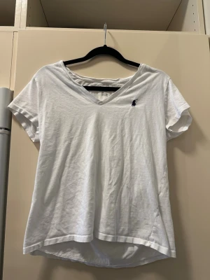 Vit v-ringad t-shirt från Polo Ralph Lauren - Vit t-shirt från Polo Ralph Lauren med v-ringning och korta ärmar. Klassisk broderad logga i marinblått på bröstet. Tillverkad i mjuk bomull för en skön och avslappnad passform.