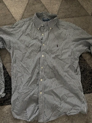 Randig skjorta från Polo Ralph Lauren - Klassisk skjorta från Polo Ralph Lauren med smala gröna och vita ränder. Skjortan har lång ärm, knappar framtill och den ikoniska broderade loggan på bröstet. Tillverkad i mjuk bomull och har en tidlös krage.