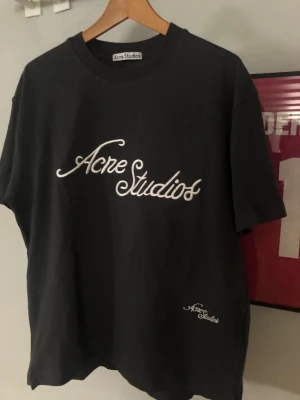 Mörkblå Acne Studios t-shirt - Snygg  t-shirt från Acne Studios med vit broderad logga framtill och mindre logga vid nederkanten. Klassisk passform och rund halsringning. Perfekt till jeans eller shorts för en clean och stilren look.