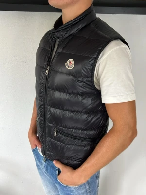 Moncler gui väst - Snygg svart dunväst från Moncler med hög krage och dragkedja framtill. Västen har två fickor med dragkedja och Moncler-logga på bröstet. Perfekt att slänga över en hoodie eller t-shirt för en clean och modern look.