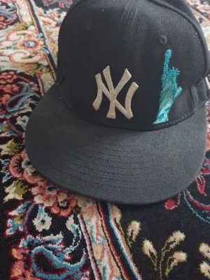 Svart New York Yankees keps - Säljer en svart New York Yankees keps från New Era med broderad NY-logga och Frihetsgudinnan framtill. Baksidan har ett äpple och MLB-logga. Klassisk rak skärm och justerbar passform. Perfekt för dig som gillar streetwear och sportig stil.
