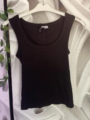 Svart ribbad linnetopp - En svart ribbad linnetopp med breda axelband och rundad halsringning. Toppen är ärmlös och har en enkel, stilren design som passar perfekt till jeans eller kjol. Materialet är stretchigt och bekvämt, vilket gör den lätt att bära hela dagen.