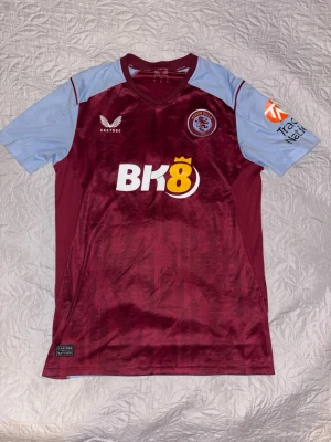Aston Villa matchtröja - Säljer en officiell Aston Villa matchtröja från Castore i vinrött med ljusblå ärmar. Tröjan har klubbmärke på bröstet, BK8 sponsortryck och Trade Nation-logga på ärmen. Tillverkad i lätt och ventilerande material, perfekt för fotboll eller träning. Köpt från Aston villas egna hemsida, äkta produkt.