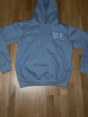 Ljusblå hoodie - Ljusblå hoodie med huva och magficka. På bröstet finns ett vitt tryck med bokstäverna 'BL' och på ryggen står det 'Benlous' i samma stil. Hoodien har ribbade muddar vid ärmslut och nederkant. Perfekt för en chill och avslappnad stil.