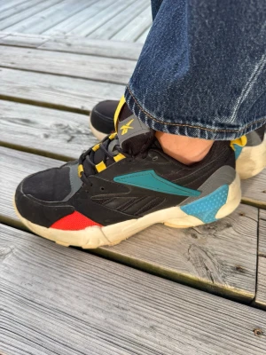 Färgglada sneakers från Reebok med retrostil - Snygga sneakers i svart med detaljer i rött, blått, gult och grått. Skorna har en chunky vit sula och coola färgblock som ger en retrovibe. Materialet är en mix av textil och syntet. Perfekta för dig som vill sticka ut med din stil.
