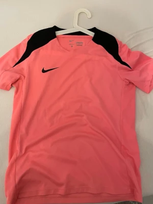 Rosa sporttröja från Nike - Snygg rosa sporttröja från Nike med svarta detaljer på axlarna och svart Nike-logga på bröstet. Tröjan har rund halsringning och är tillverkad i ett lätt och ventilerande material, perfekt för träning eller match.
