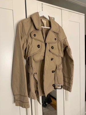 Beige trenchcoat med bälte och detaljer - Stilren beige trenchcoat med dubbelknäppning, bred krage och dekorativa axelklaffar. Jackan har ett matchande bälte i midjan och markerade sömmar för extra detalj. Tillverkad i ett lätt tyg, perfekt för lager-på-lager och klassisk look.