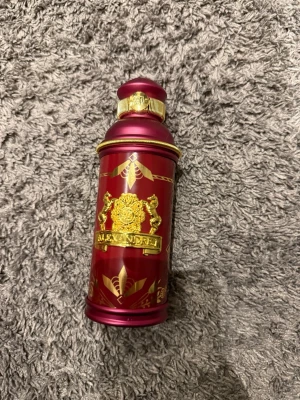 Röd parfymflaska med gulddetaljer - Snygg parfymflaska i metall med djup röd färg och eleganta gulddetaljer. Flaskan har ett dekorativt mönster med lejon och emblem samt guldfärgad ring runt halsen. Perfekt som en lyxig detalj på parfymhyllan.