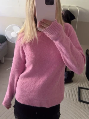 Ljusrosa fluffig stickad tröja - Mysig ljusrosa stickad tröja med rund halsringning och långa ärmar. Tröjan har en fluffig, mjuk struktur och ribbstickade muddar vid ärmslut och nederkant. Perfekt för att hålla värmen under kyliga dagar.
