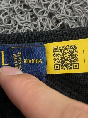 Svart kabelstickad tröja Polo Ralph Lauren - Svart kabelstickad tröja från Polo Ralph Lauren med lila broderad logga på bröstet. Tröjan har rund halsringning, långa ärmar och ribbade muddar. Perfekt för en stilren och klassisk look.