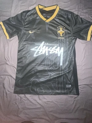 Brazilian special kits Nike x stussy black - Snygg svart Brasilien-tröja i samarbete mellan Nike och Stüssy. Tröjan har gula detaljer vid krage och ärmslut, Brasilien-emblem på bröstet och Stüssy-logga tryckt över magen. Tillverkad i lätt och ventilerande material, perfekt för sport eller streetwear.