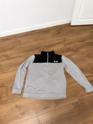 Grå ziptröja från Under Armour - Snygg grå ziptröja från Under Armour med svart panel upptill och logga på bröstet. Tröjan har hel dragkedja, hög krage och långa ärmar. Perfekt för träning eller chill dagar. Materialet är mjukt och stretchigt för extra komfort.