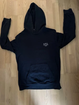 Svart hoodie från A.P.C. - Snygg svart hoodie från A.P.C. med vit brodyr på bröstet där det står 'A.P.C. RUE MADAME PARIS'. Klassisk känguruficka framtill och stor huva. Tillverkad i mjukt material som känns skönt mot huden. Perfekt för en clean och stilren look.